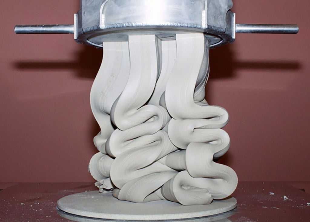 Soft white clay extruding from a metal press, folding into thick, flowing ribbons that coil into an organic sculptural form against a muted terracotta background.