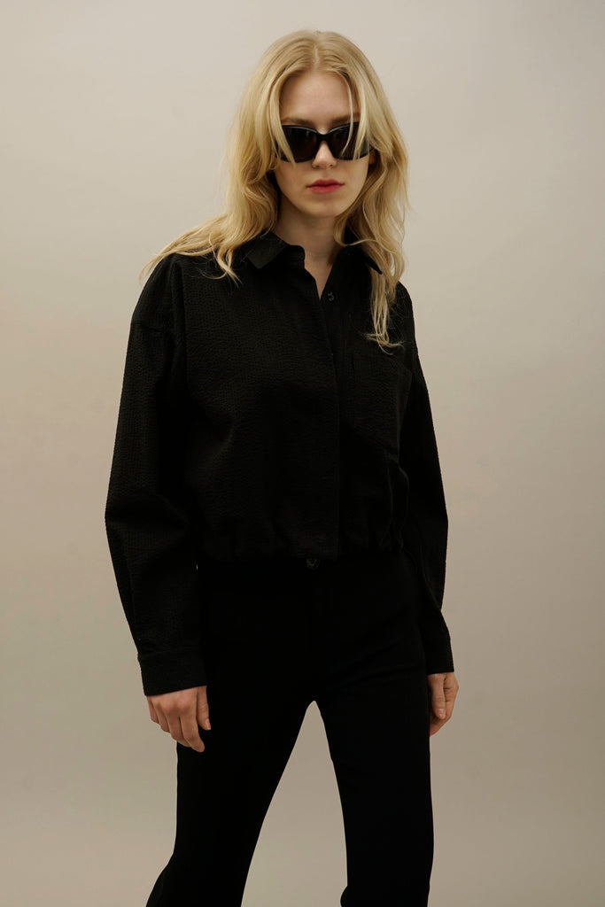Woman wearing black textured button-up shirt and slim black pants, styled with sunglasses