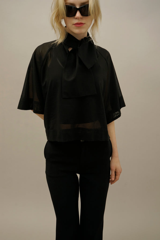 Woman wearing a black sheer blouse with a large bow tie, paired with black pants and stylish sunglasses, exemplifying modern fashion.