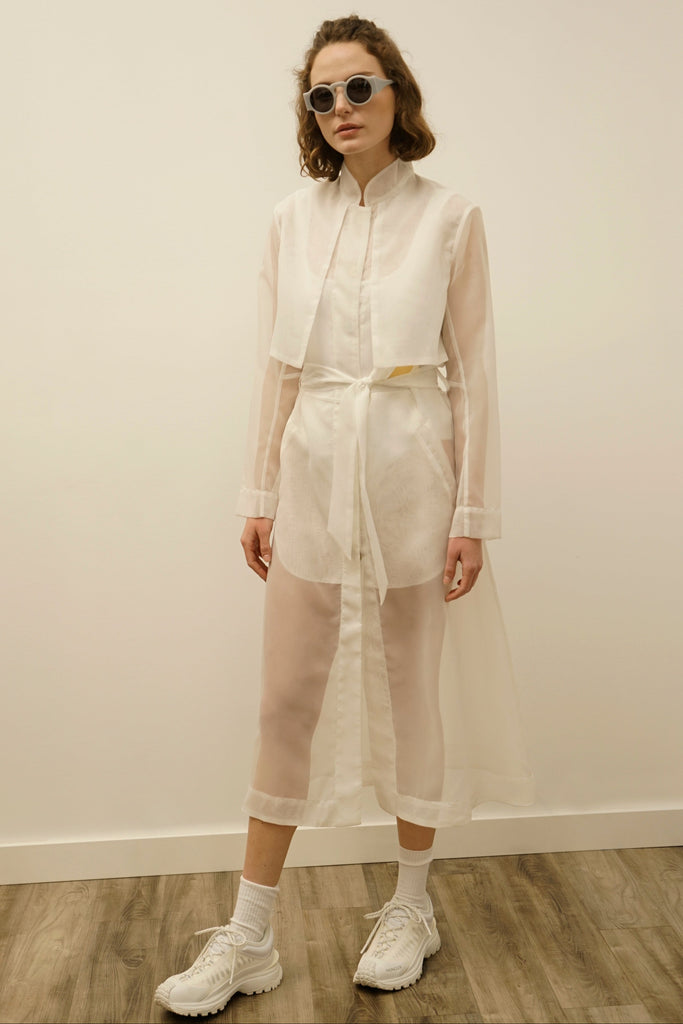 Fashion model showcasing a transparent white trench dress with a belt by NINObrand, paired with sporty white sneakers and stylish sunglasses, set against a minimalist background.