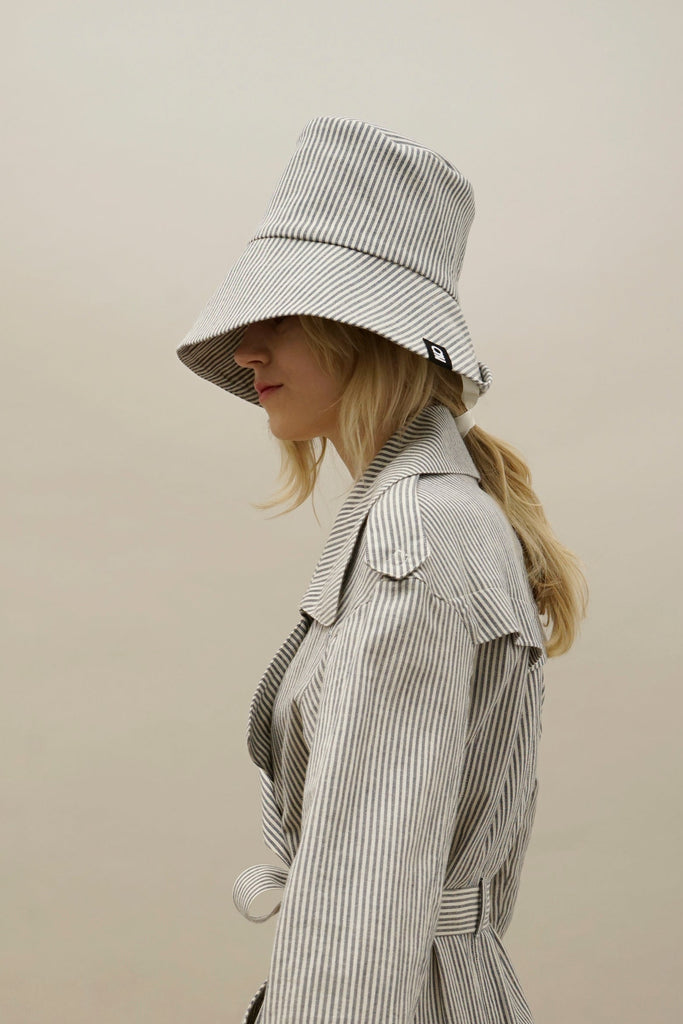 Side view of model in a NINObrand pinstripe spring coat and large brim bucket hat, showcasing modern neutral-toned outfit