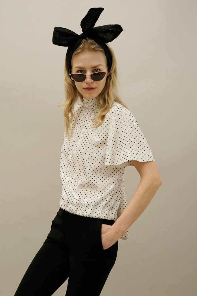 Woman wearing white polka dot blouse with high neckline, black pants, oversized black bow headband, and small sunglasses.